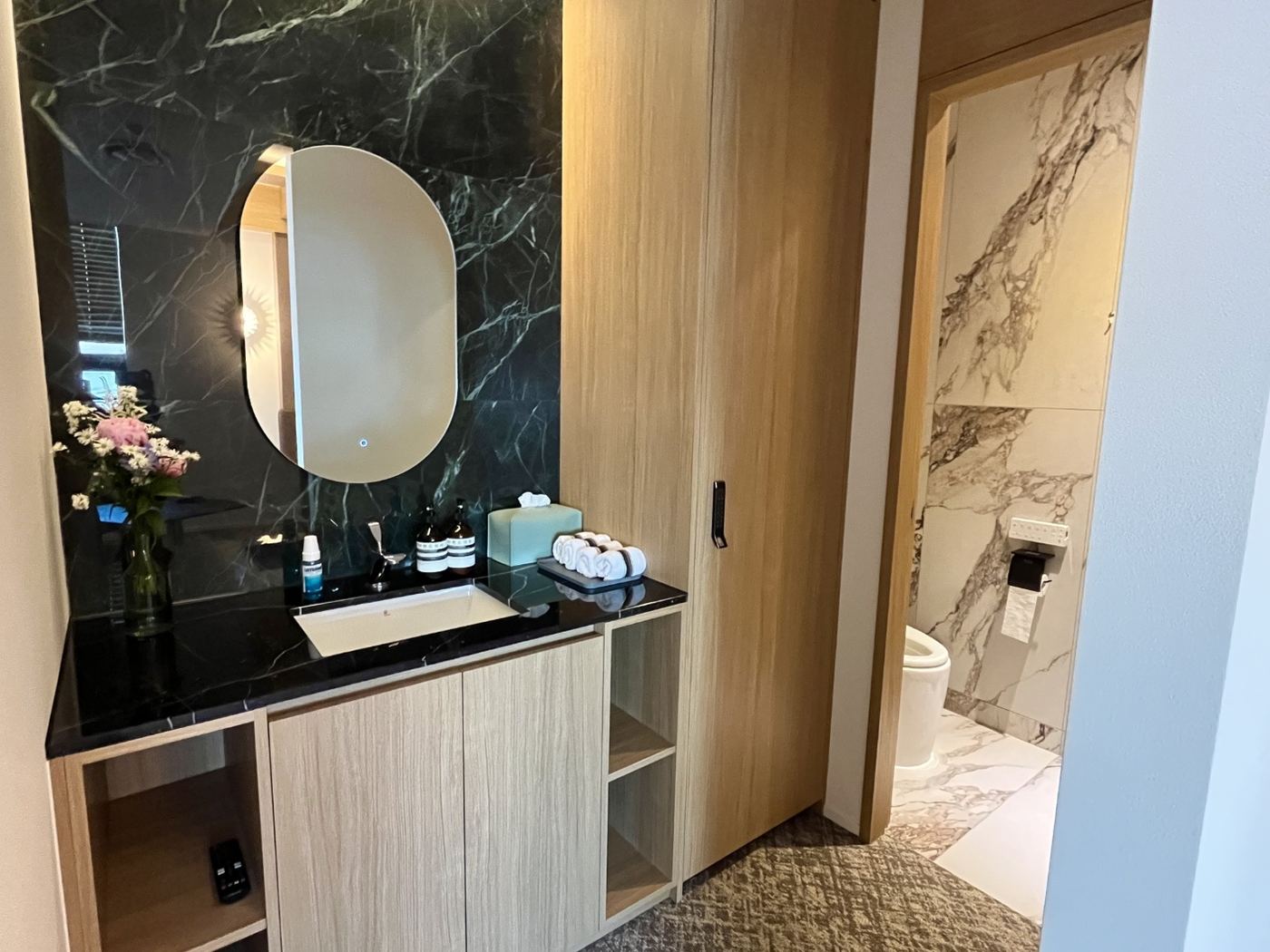 Black marble vanity corner in an EHL Bio patient suite with an oval LED mirror, skincare amenities, rolled towels and a timber door opening to a white-marble en-suite bathroom