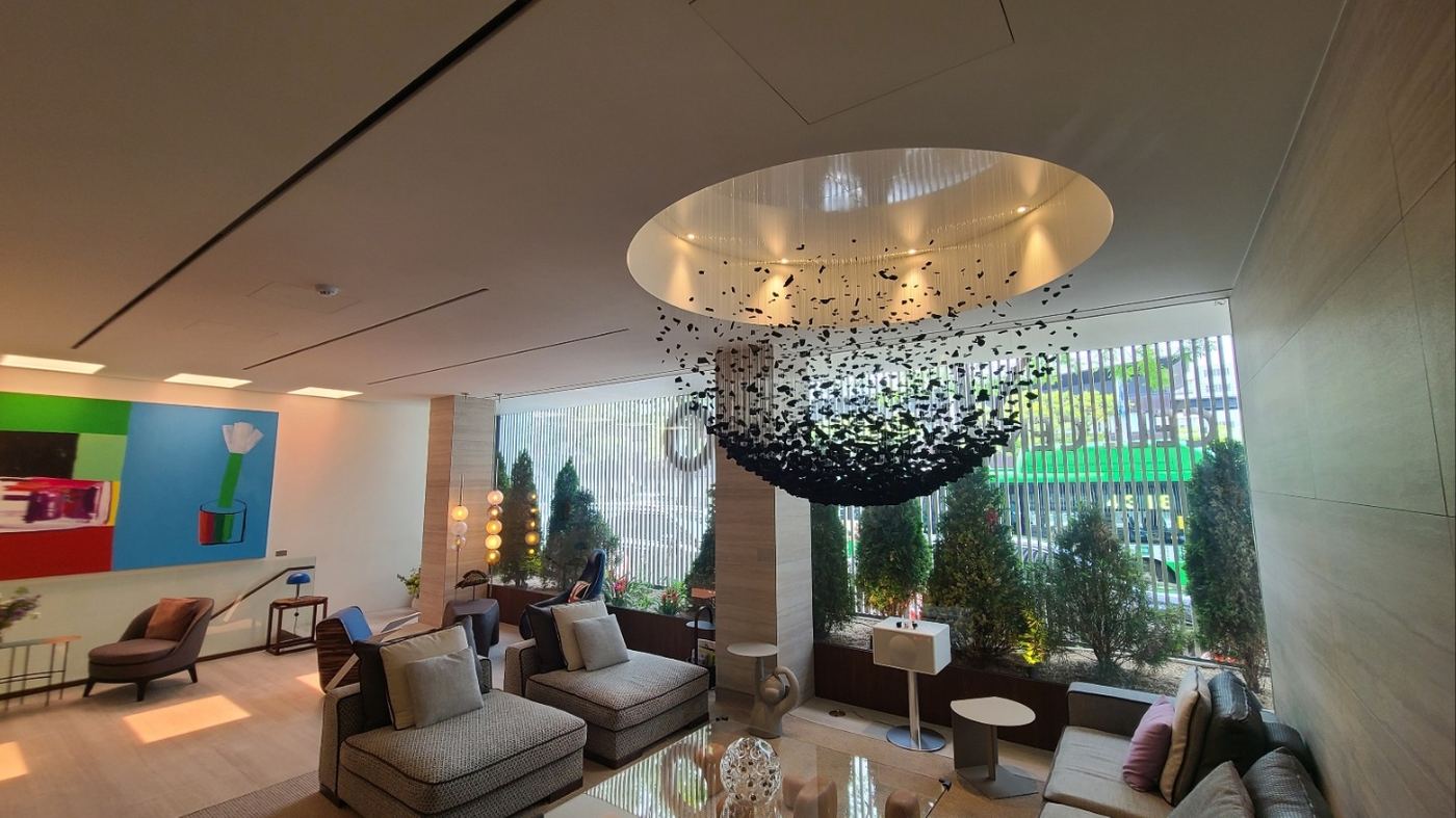 EHL Bio waiting lounge in cream tones with a sculptural mobile hanging from a circular ceiling recess, woven armchairs, a bright modern painting and a row of potted conifers along the tall windows