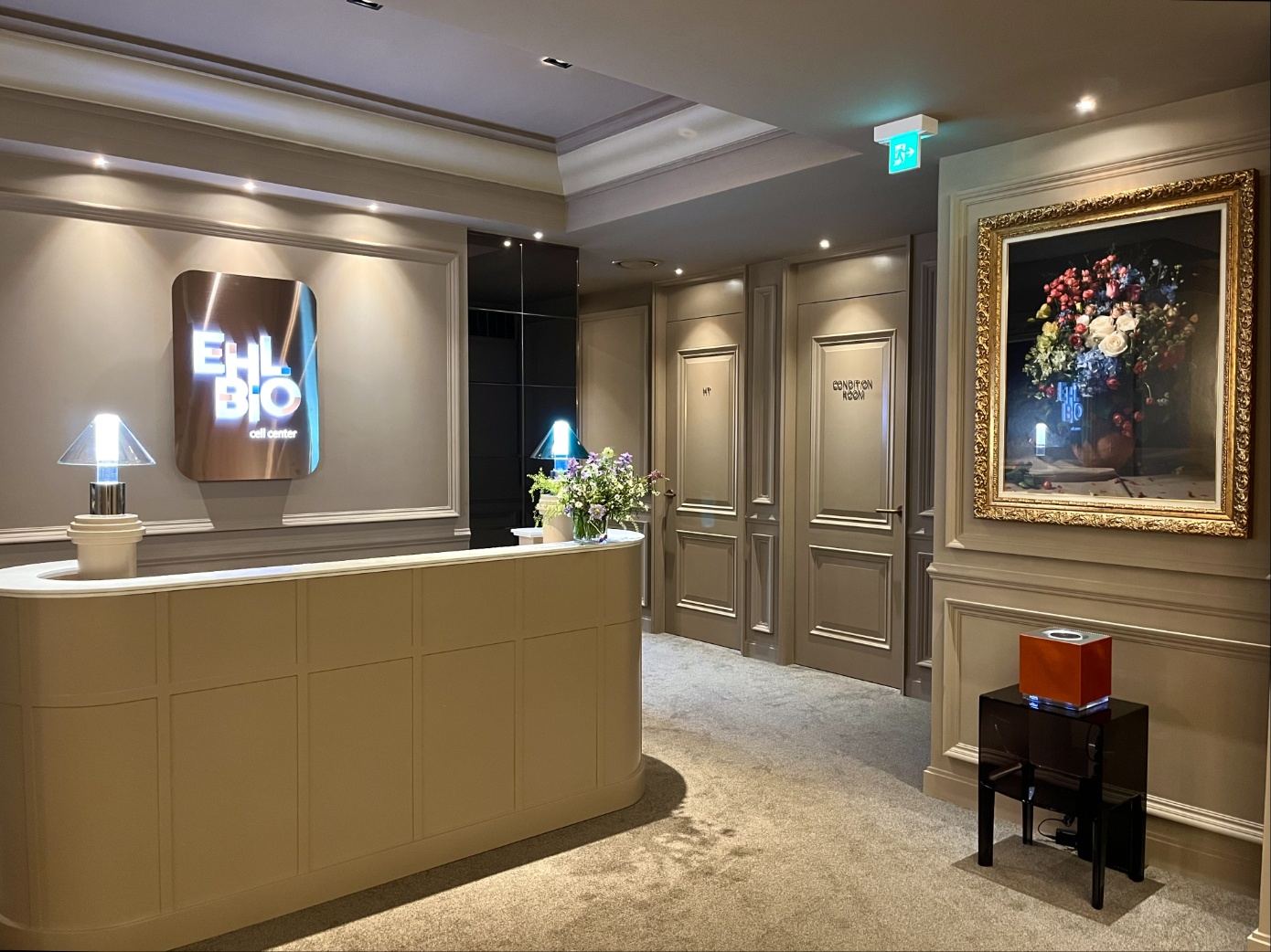 Curved beige reception desk at the EHL Bio Cell Center with an illuminated EHL BIO cell center logo on a classically panelled wall, table lamps, fresh flowers and a gilt-framed floral painting to the right