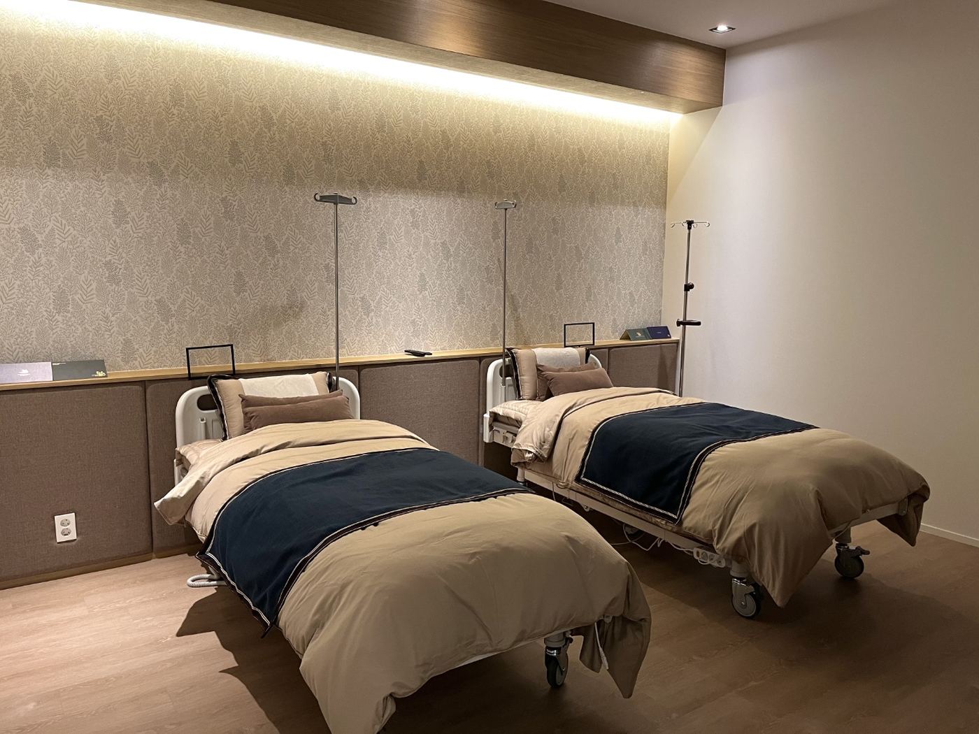 Twin-bed EHL Bio recovery room with hospital beds under beige duvets and navy runners, IV poles beside each bed, and a leaf-patterned wall lit by concealed lighting under a timber soffit