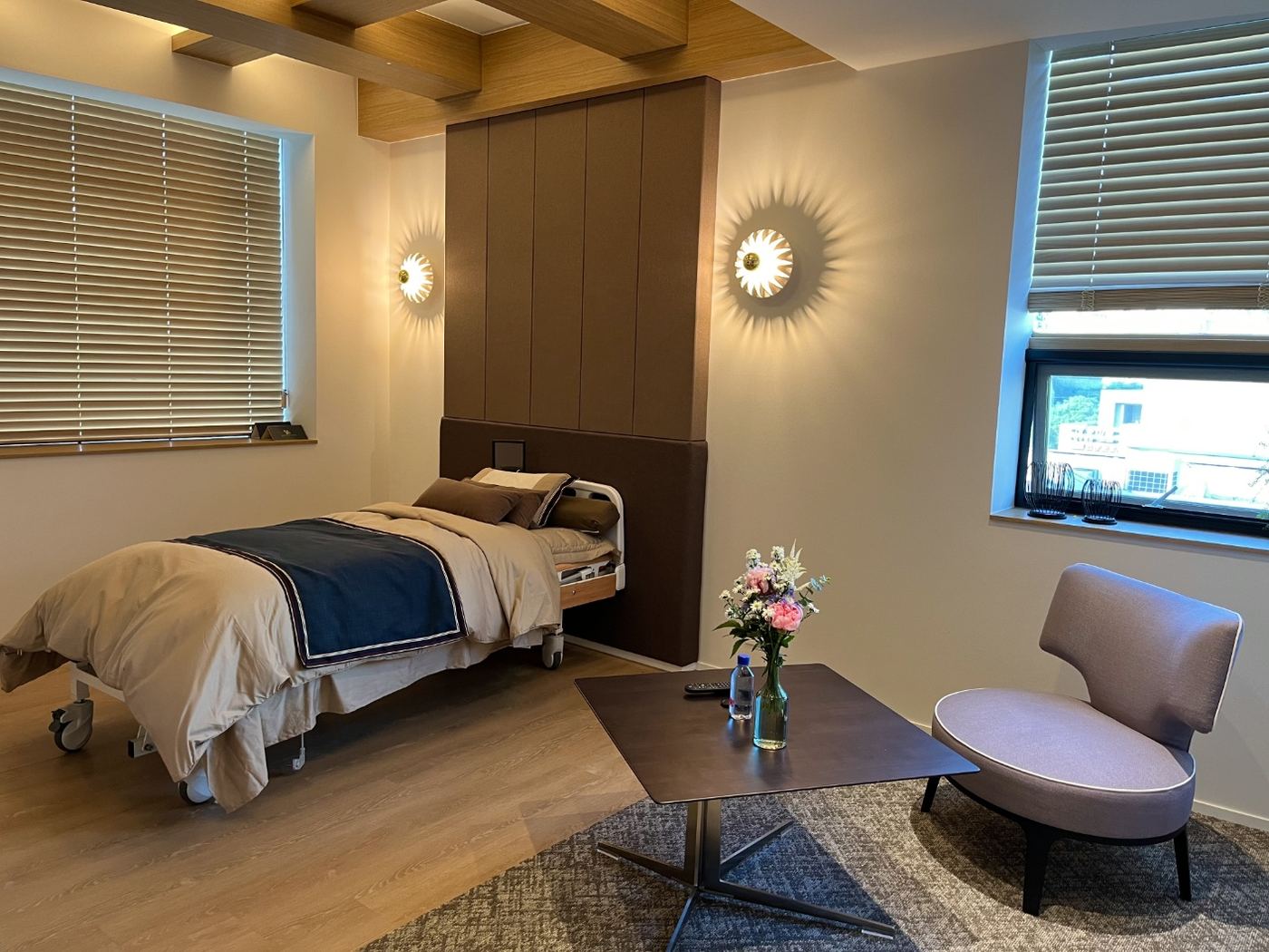 Private EHL Bio infusion suite with an adjustable hospital bed dressed in beige and navy linen, timber-beamed ceiling, floral wall sconces, a low table and an upholstered chair for a companion, lit by windows on two sides